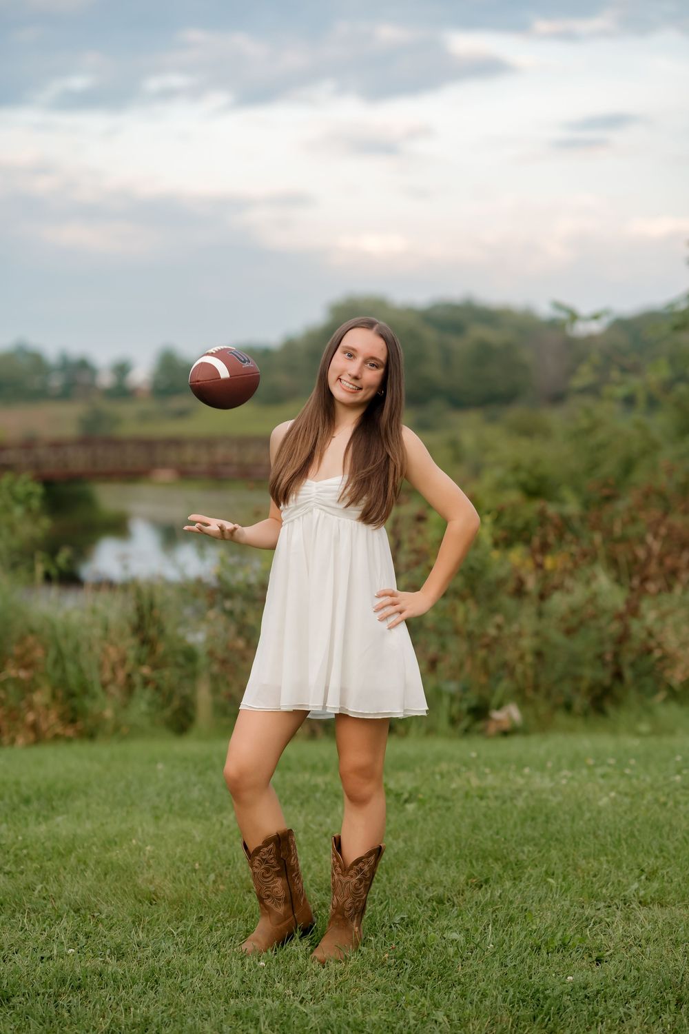 Young woman in white dress and cowboy boots tossing a football outdoors near a scenic bridge.