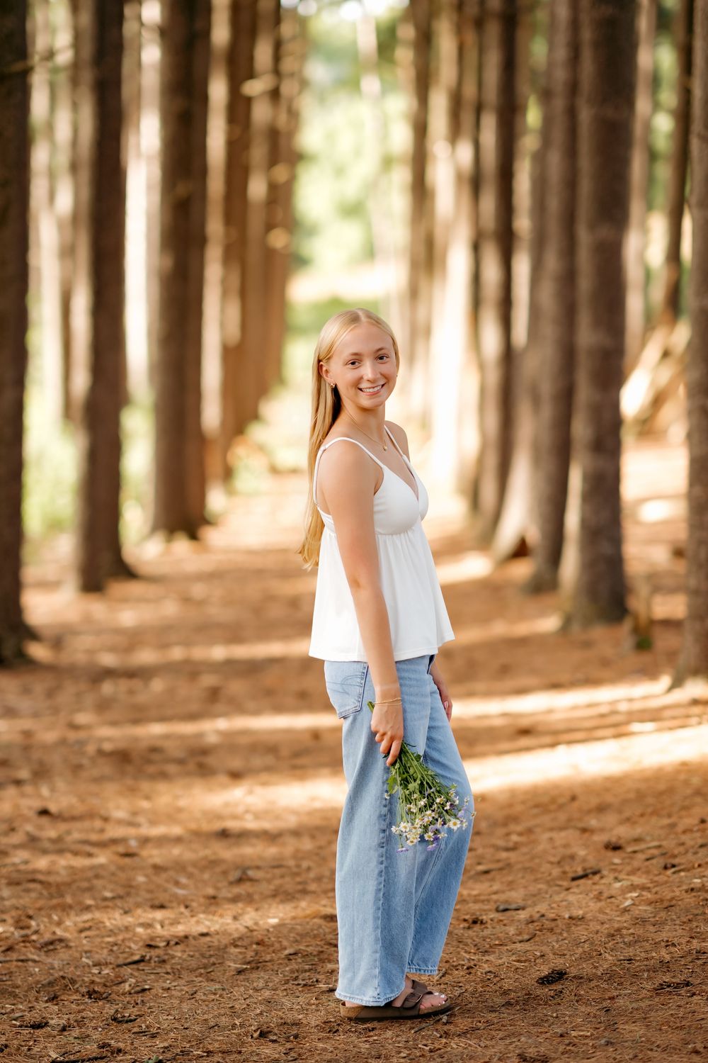 Smiling blonde teen in white top and jeans holds wildflowers on a pine forest path.
