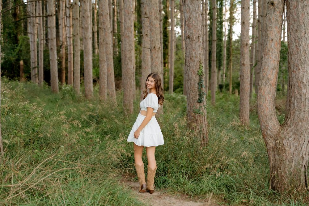 Young woman in white dress and brown boots standing on a dirt path among tall pine trees in a lush forest.