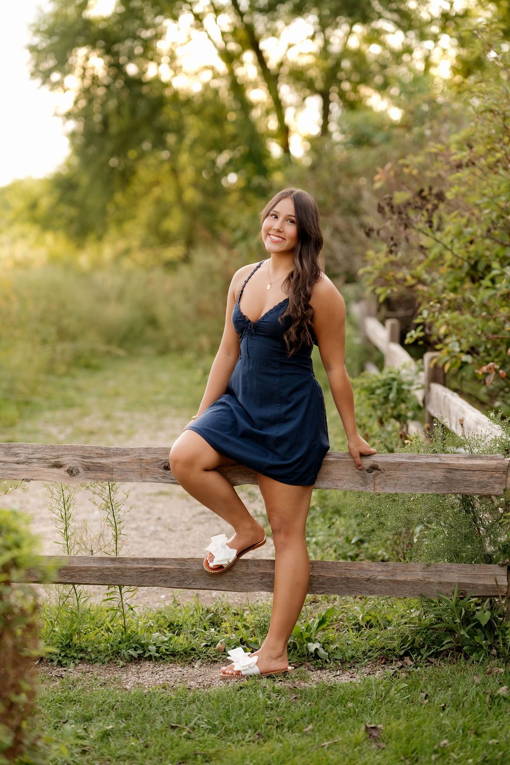 Young woman in navy blue dress smiling while leaning on rustic wooden fence in lush green outdoor setting.