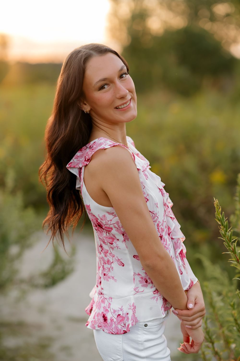 Smiling woman in floral pink top and white jeans posing outdoors at golden hour sunset.
