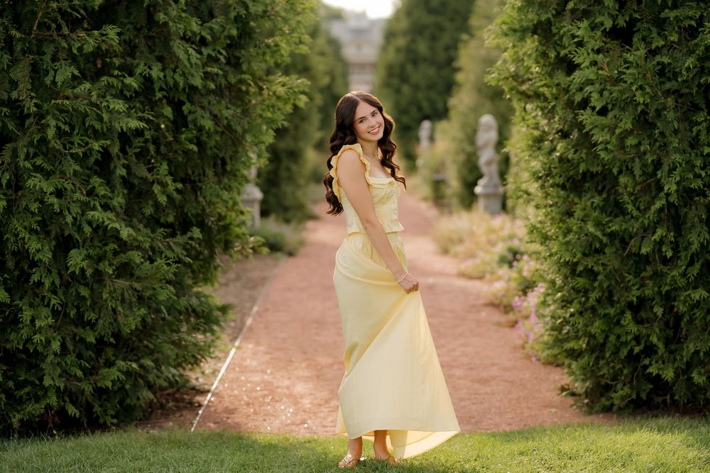 Young woman in elegant yellow gown poses gracefully in a lush garden pathway lined with tall green hedges.