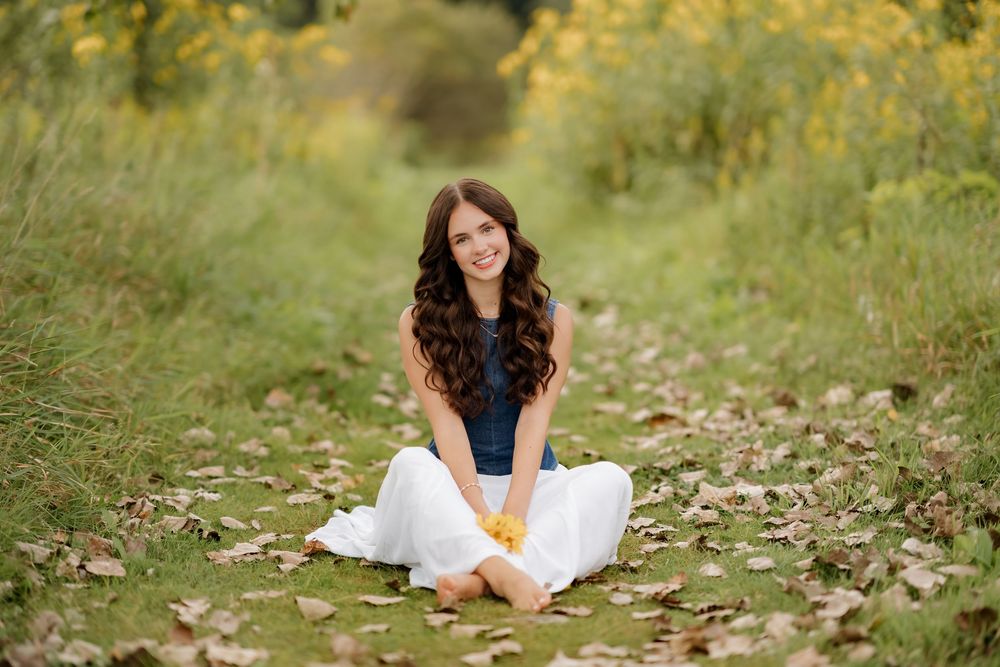 A smiling young woman with curly hair sits cross-legged on a leaf-covered path in a lush green outdoor setting.