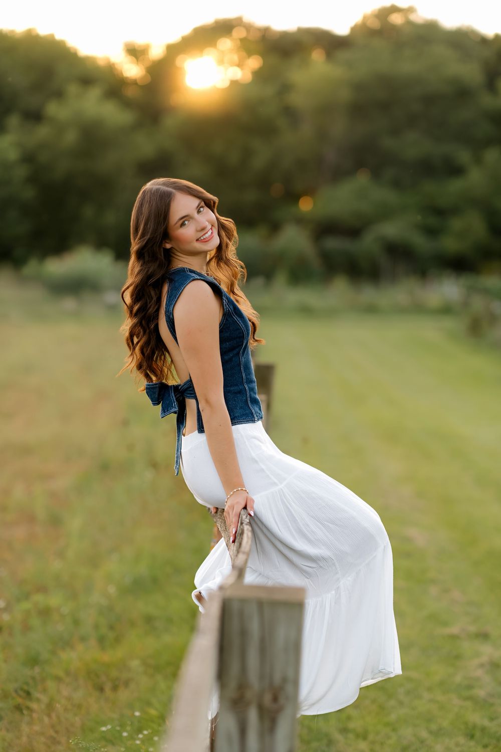 Young woman in denim top and white skirt leaning on wooden fence at golden hour sunset in green field.
