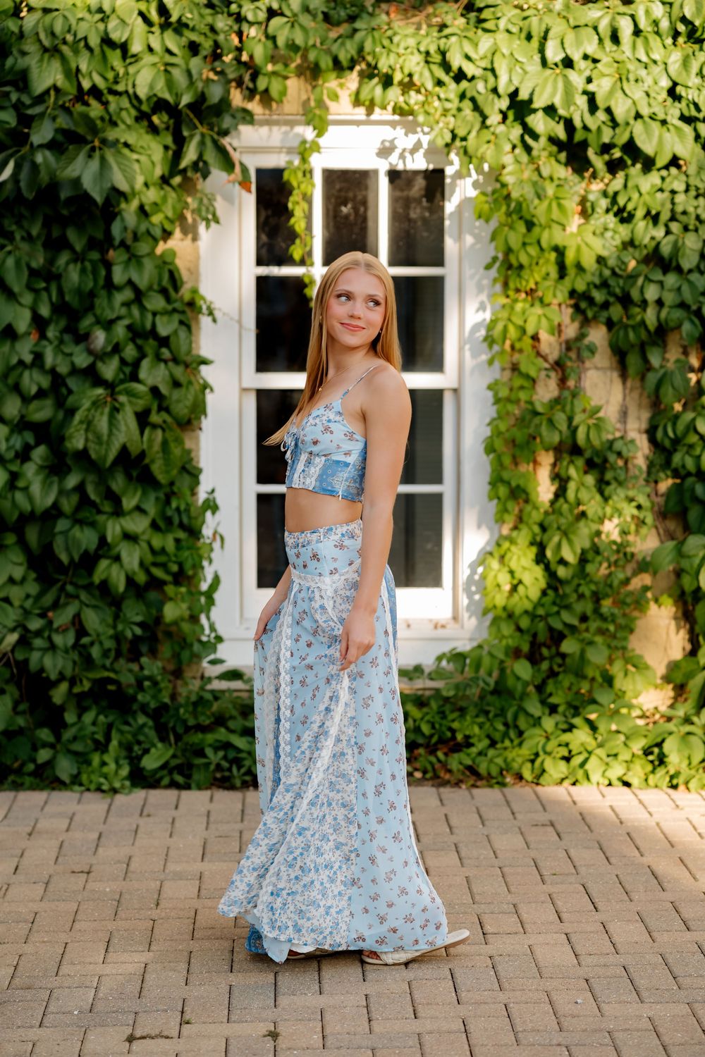 Young woman in blue floral two-piece maxi dress poses outdoors by ivy-covered wall and white window.