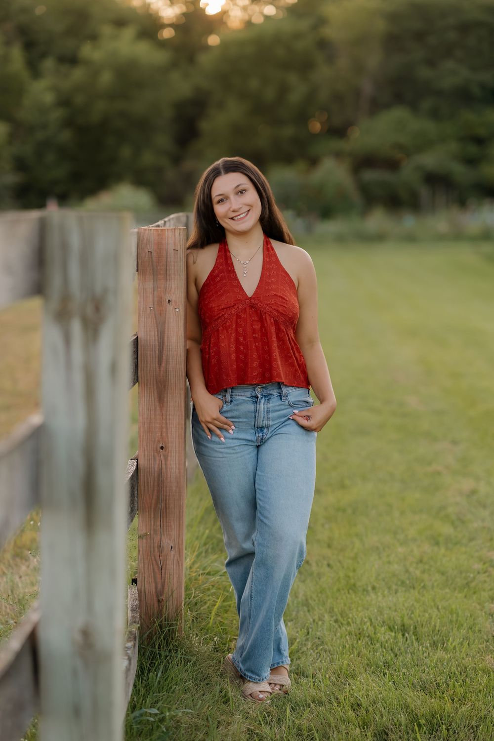Young woman in red halter top and jeans leaning against wooden fence in green field at golden hour.