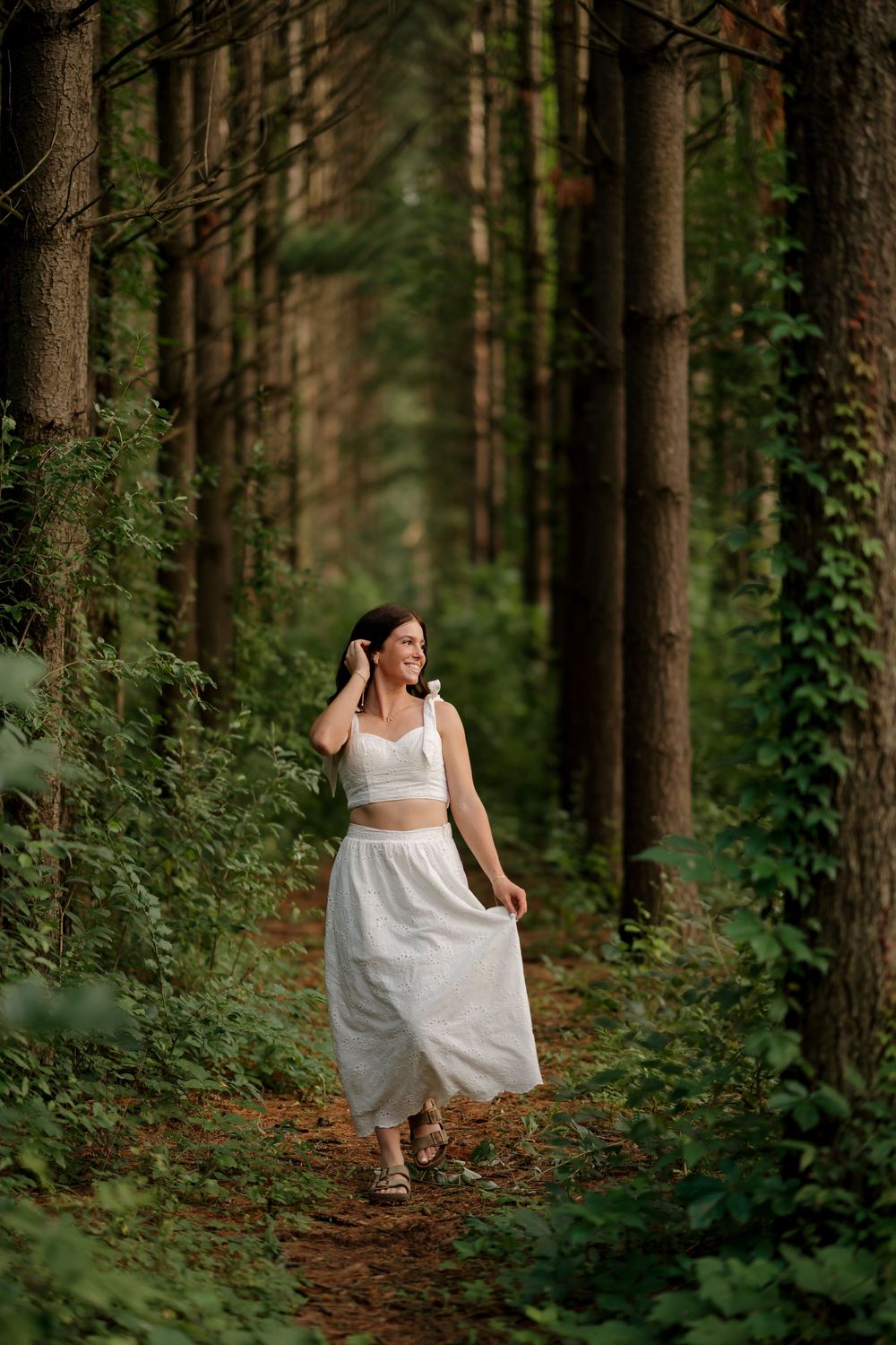 Woman in white crop top and skirt walking on a forest path surrounded by tall trees and lush greenery.