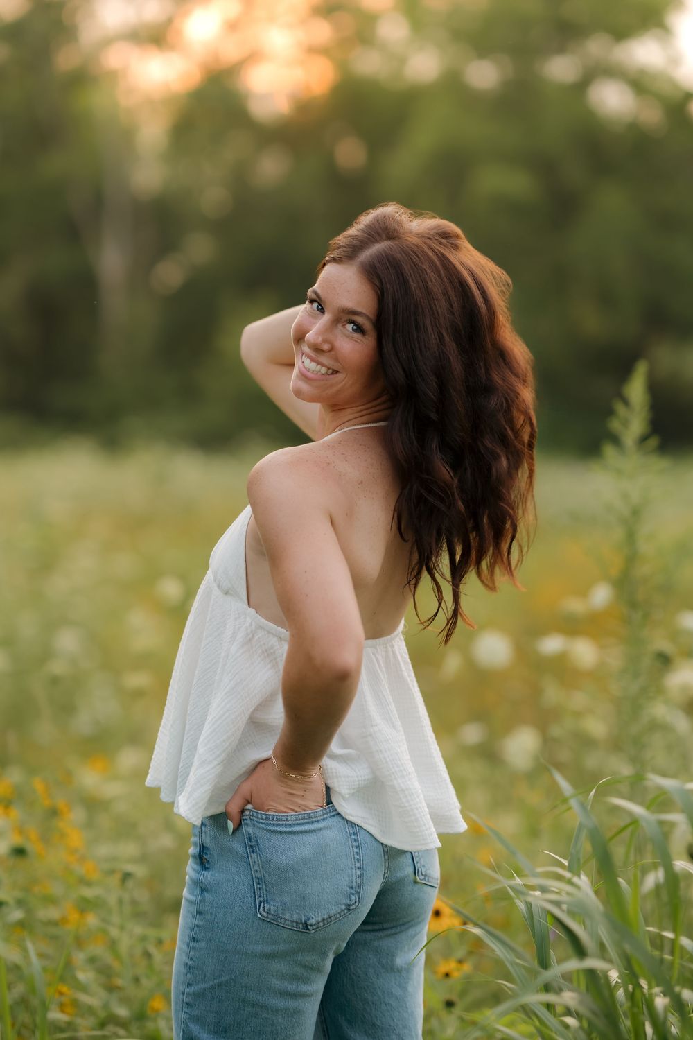 Young woman in white top and jeans smiling over shoulder in a sunny wildflower field at golden hour.