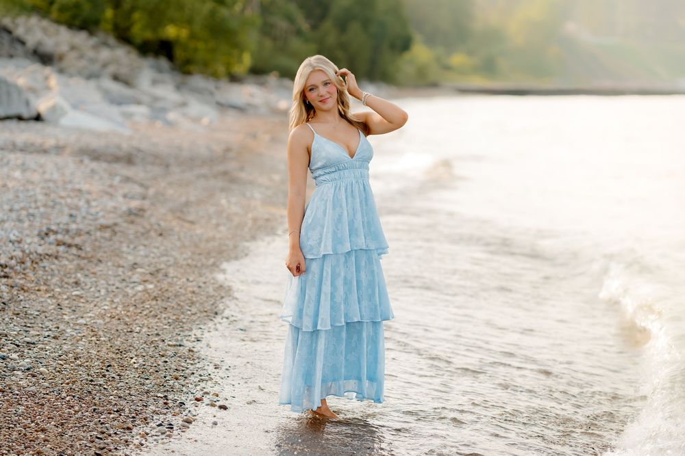 A blonde woman in a light blue tiered maxi dress walks barefoot along a rocky beach shoreline at sunset.
