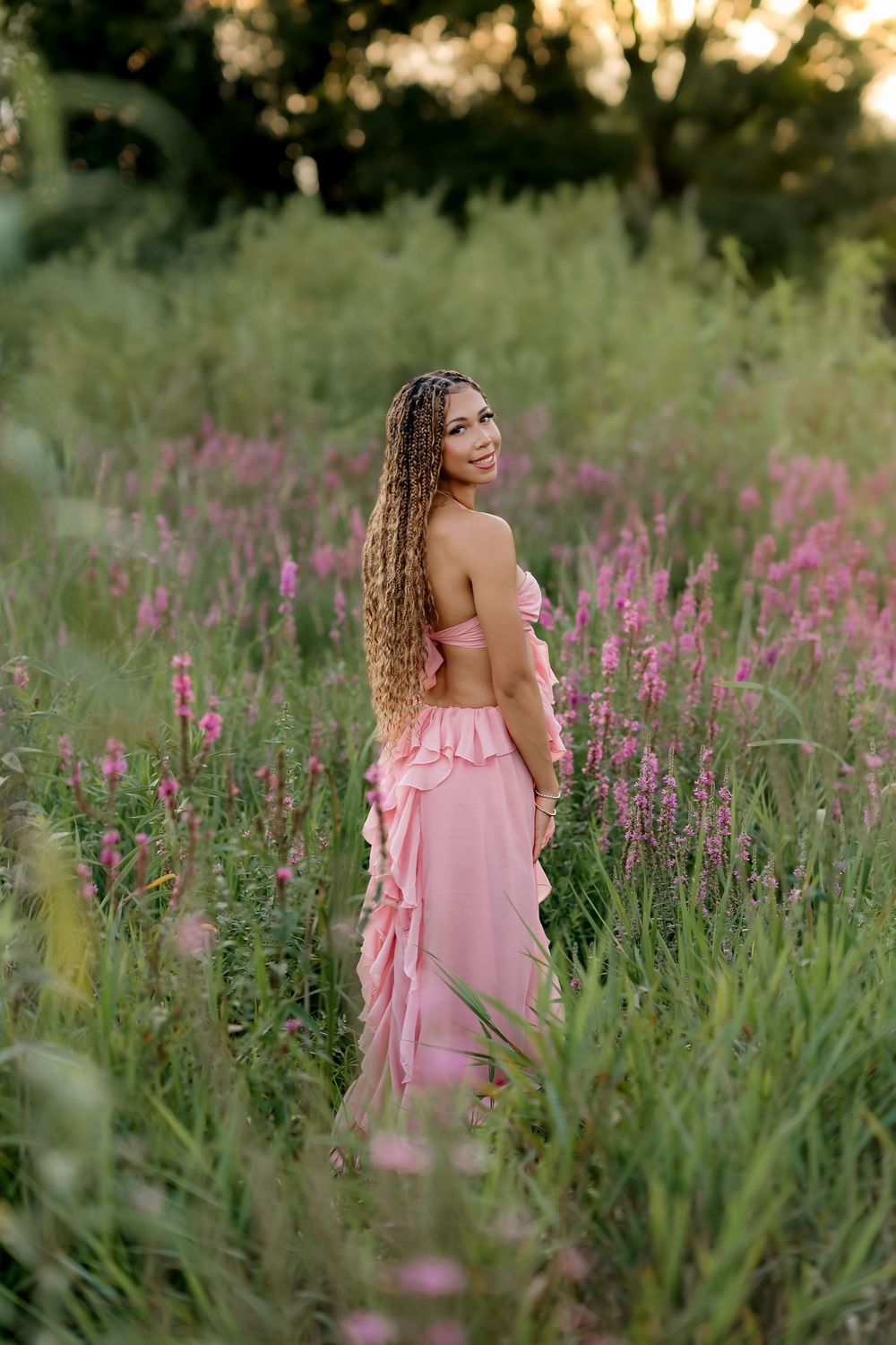 Woman in pink ruffled dress standing in wildflower field at golden hour with long curly hair.