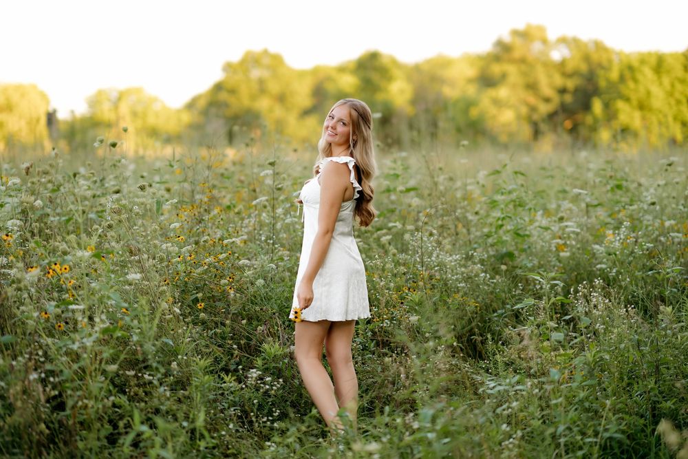Young woman in white dress standing in wildflower field during golden hour sunset photography.