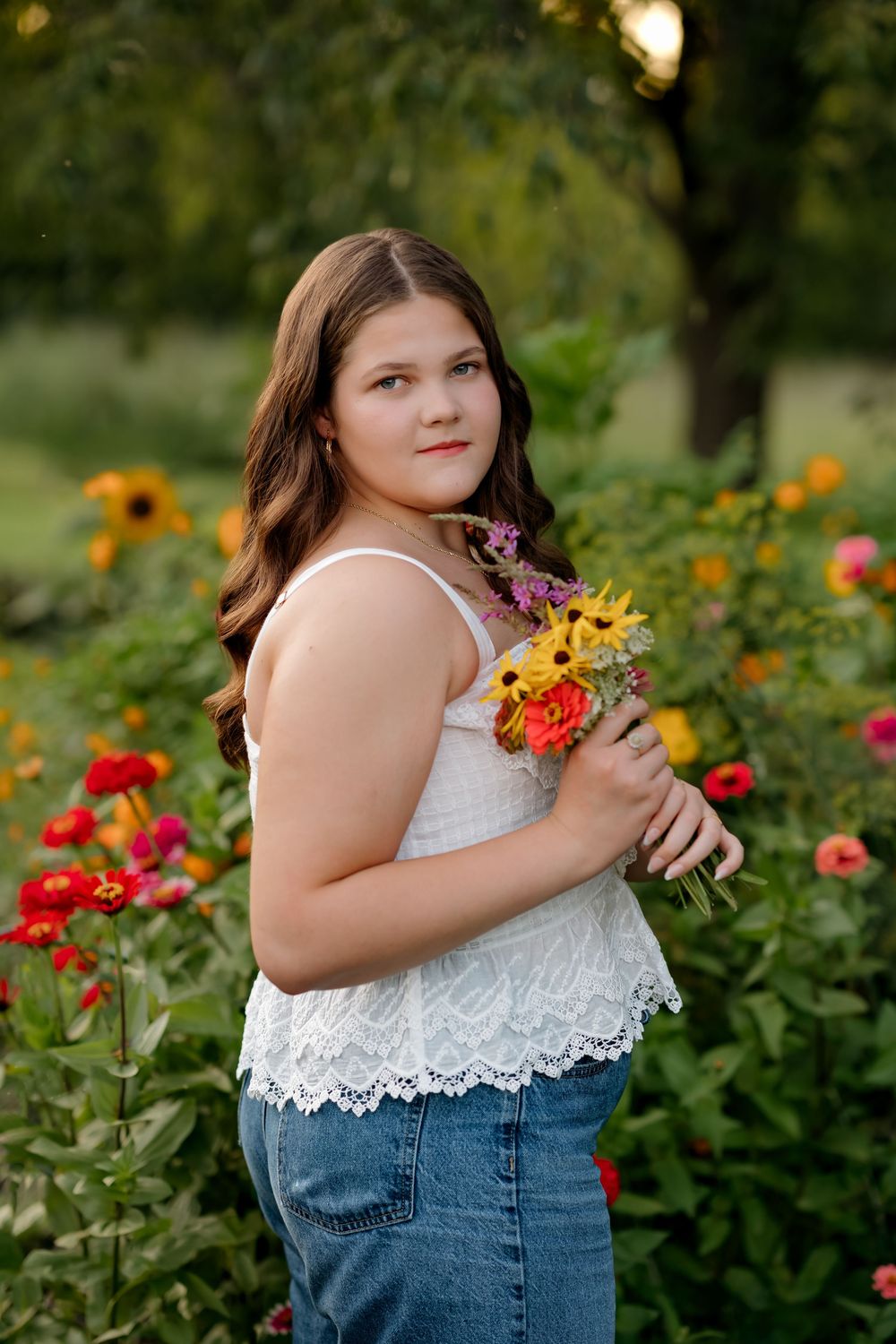 A young girl in a white lace top and jeans holds a colorful wildflower bouquet while standing in a vibrant garden.