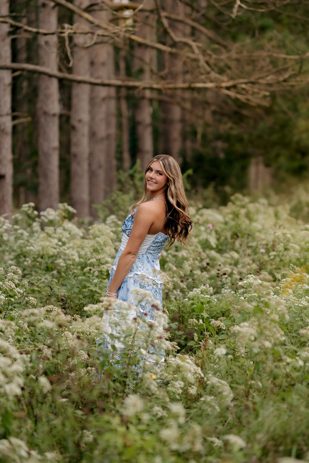 Young woman in a blue floral dress standing among white wildflowers in a lush forest setting, smiling over her shoulder.