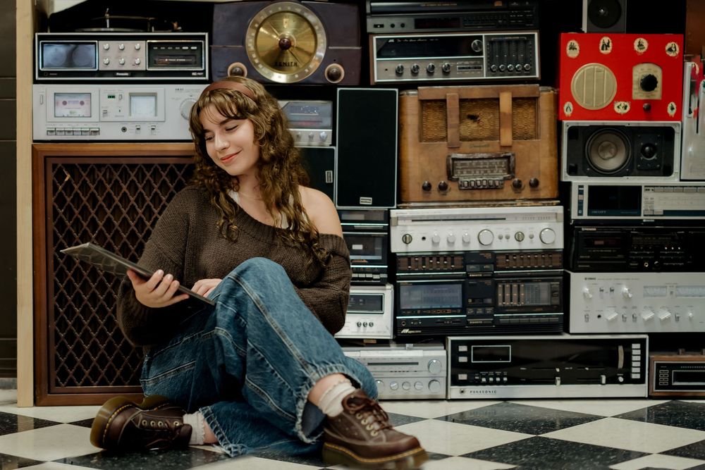 Young woman sitting on checkered floor holding tablet, surrounded by stacked vintage radios and stereos.
