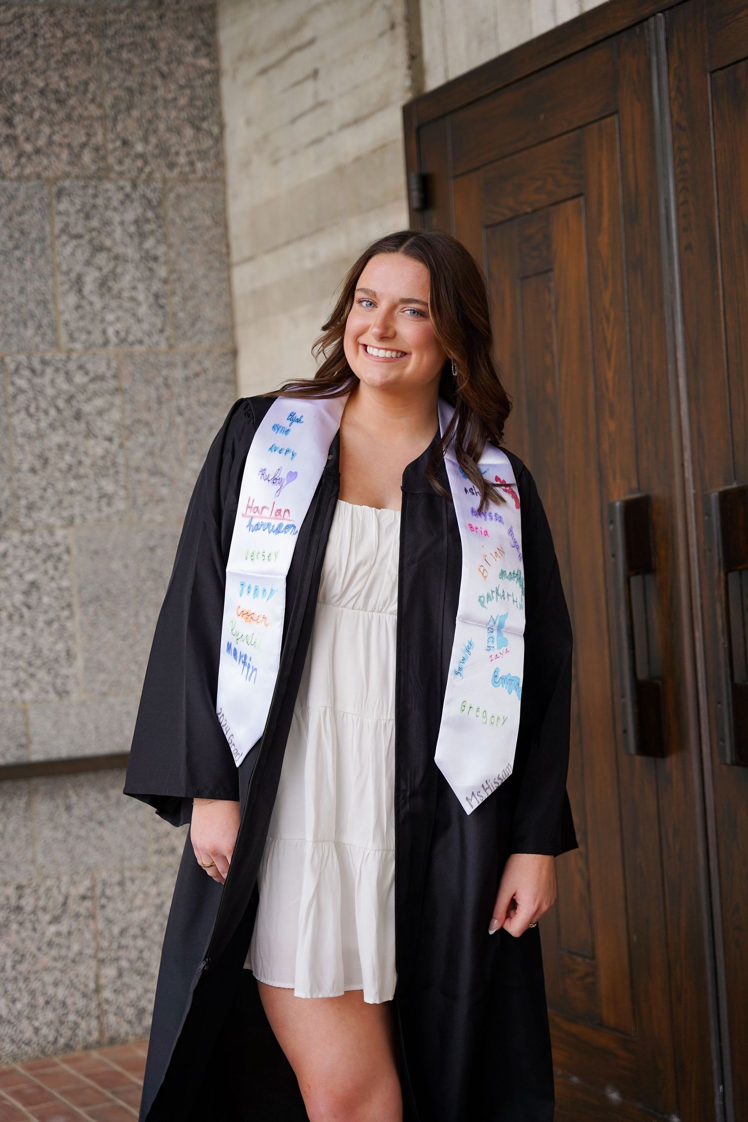 Graduate in black cap and gown with decorated sash poses by wooden door.