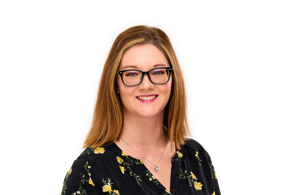 Young woman with red hair and black glasses smiling, wearing a floral top, professional headshot on white background.