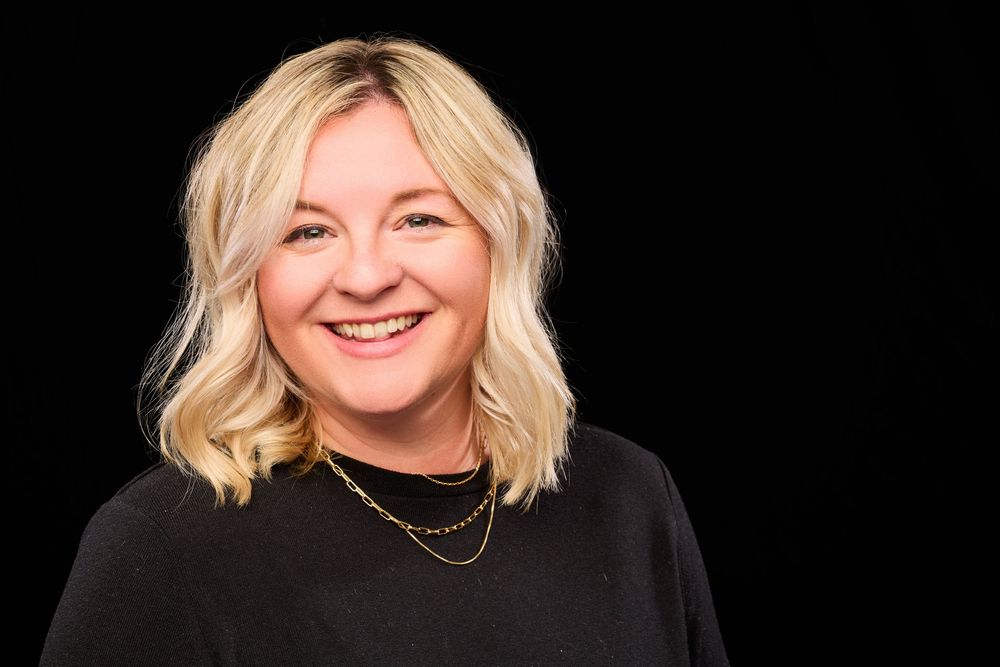 Professional headshot portrait of someone with wavy blonde hair wearing a black sweater and gold necklace against dark background.
