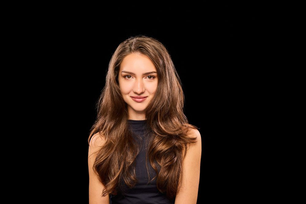 A woman with long brown hair poses in a black sleeveless top against a dark background.