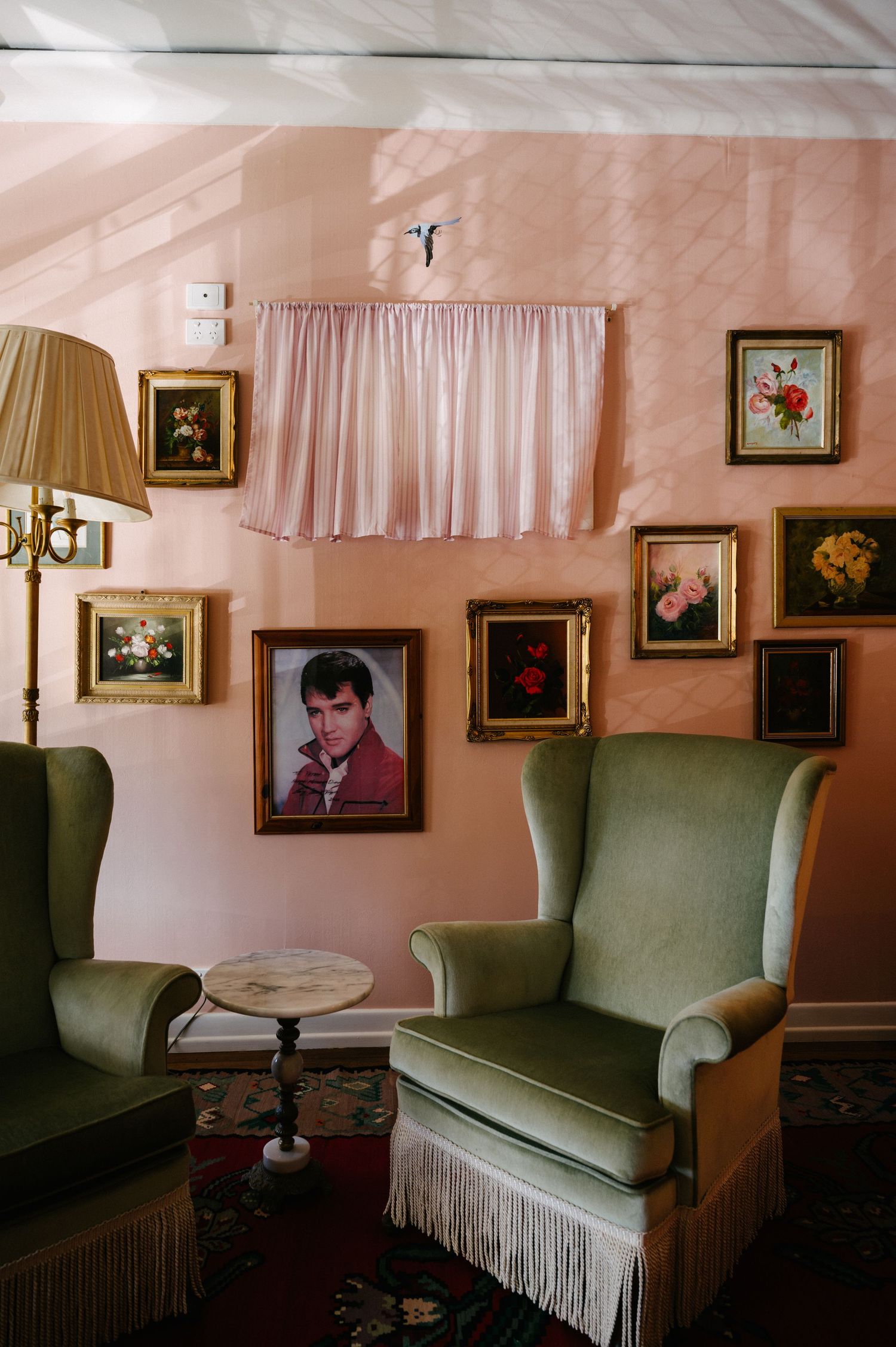 A green vintage couch with white fringe trim sits beneath framed artwork and a lamp on a pink wall in a retro interior.