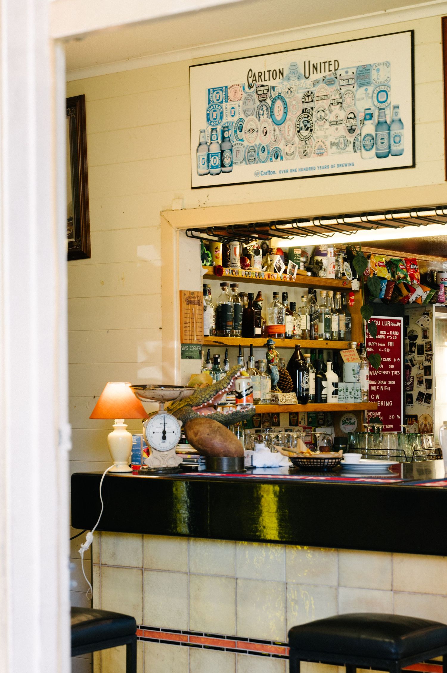 A cozy bar interior with barstools, shelves of liquor bottles, and vintage beer artwork hanging on the cream-colored wall.