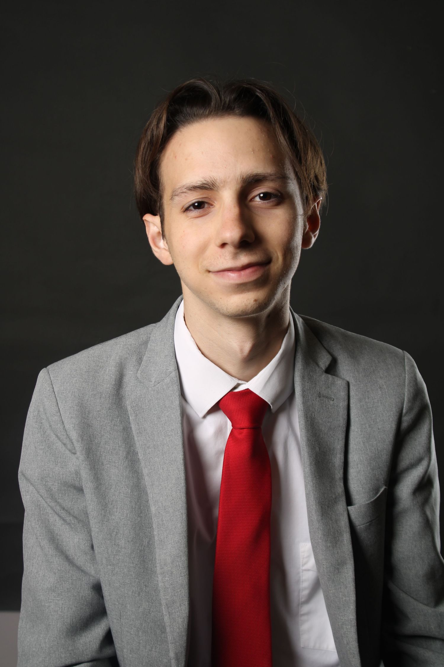 Professional headshot of a young professional in a grey suit with red tie against dark background.