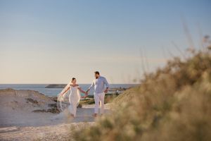 A couple in Cyprus in white outfits holds hands while walking along a scenic beach at sunset during a wedding photography shoot.