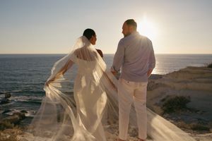 Two people in white outfits walk along a scenic beach at sunset while holding hands and gazing at the ocean views.