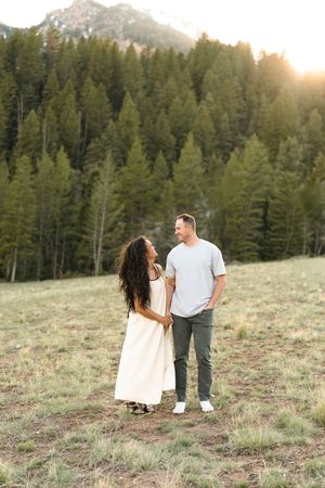 Couple holding hands in a mountain meadow at golden hour with pine trees and peaks in the background.