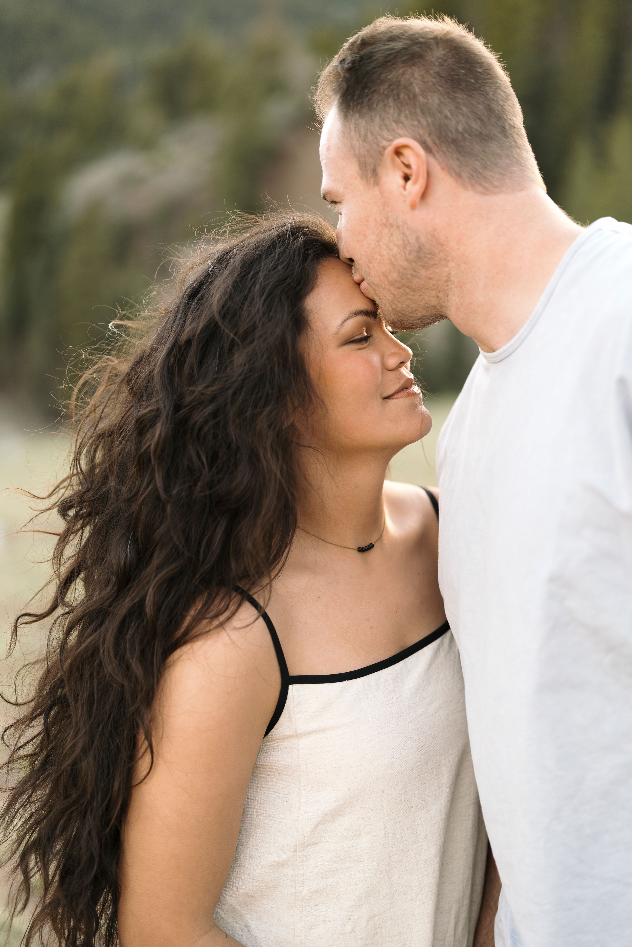 A man tenderly kisses a woman on the forehead outdoors with lush green trees in the background.
