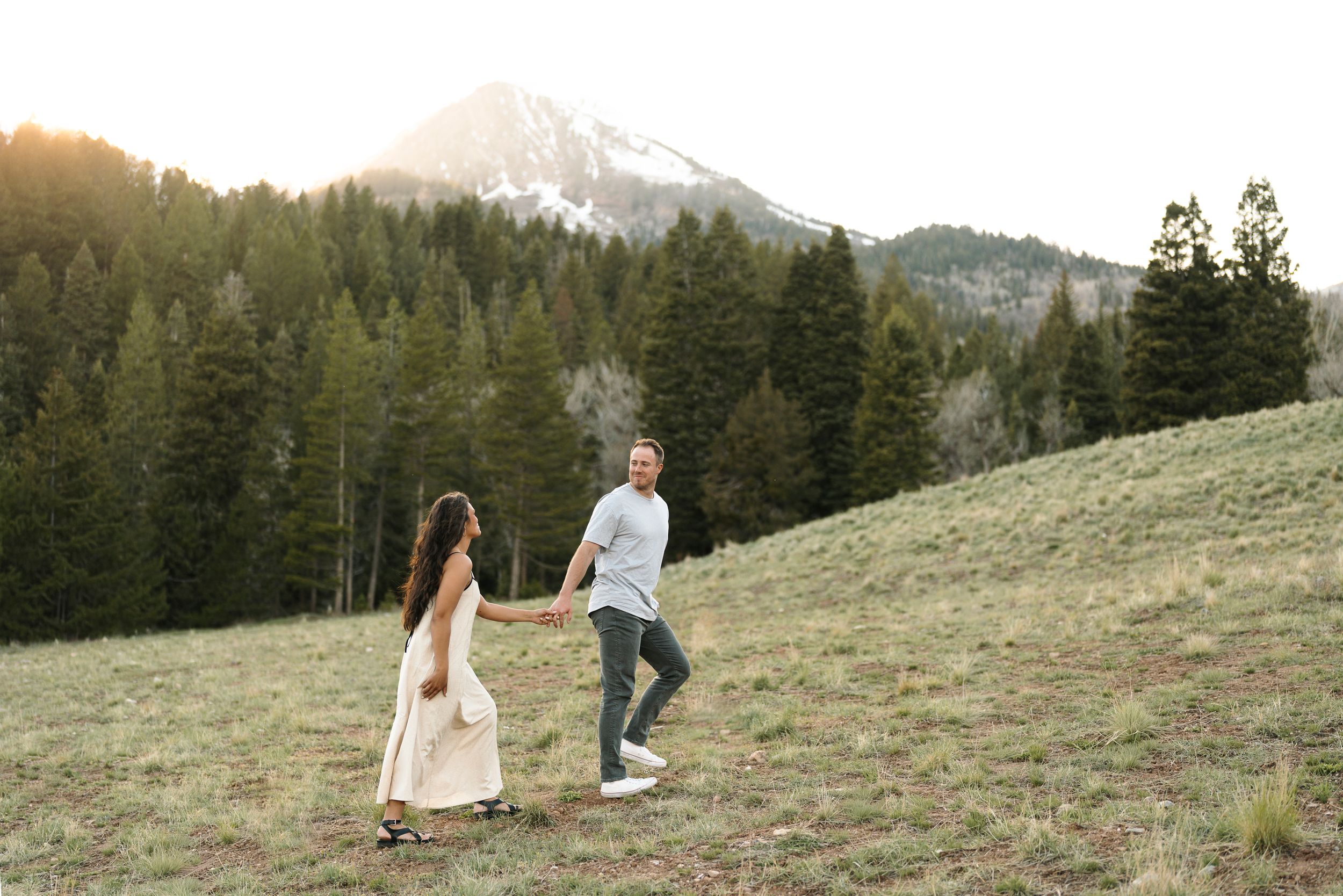 Couple holding hands walking through a mountain meadow with pine trees and snow-capped peak at golden hour.