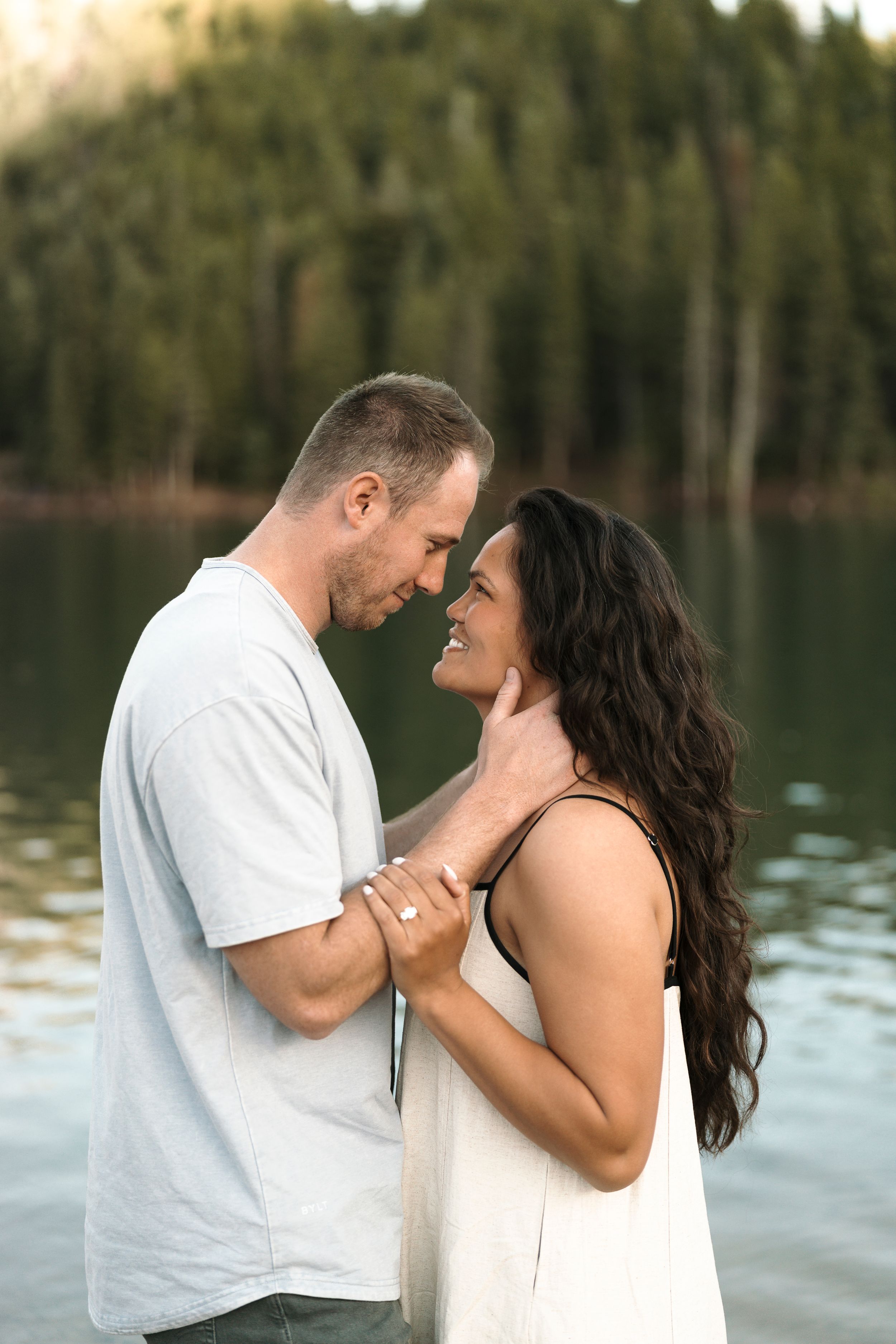 Couple sharing a romantic moment by a mountain lake, smiling and holding hands, dressed in white outfits.