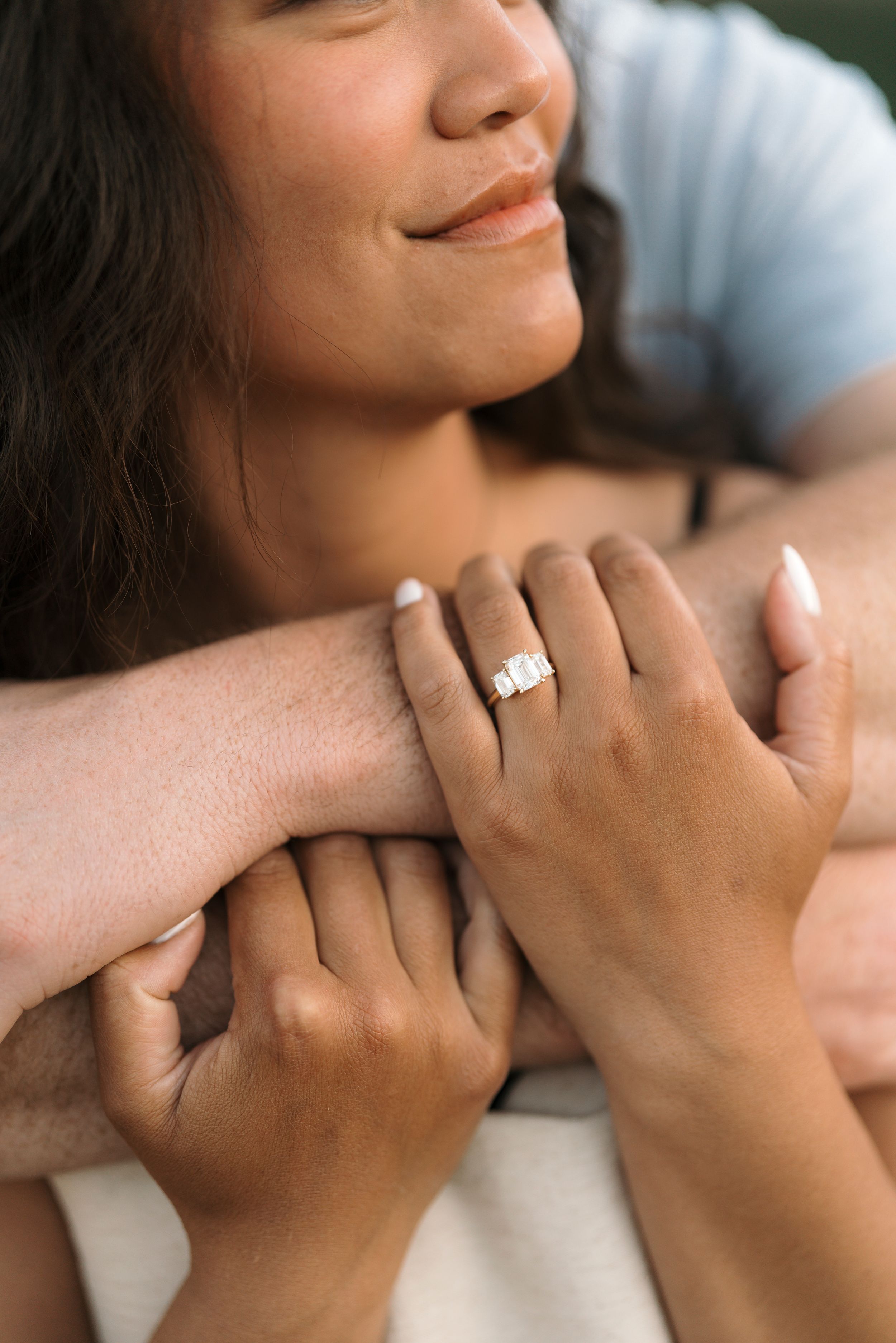 Woman smiling with engagement ring on finger, embraced by partner, close-up romantic couple photo.