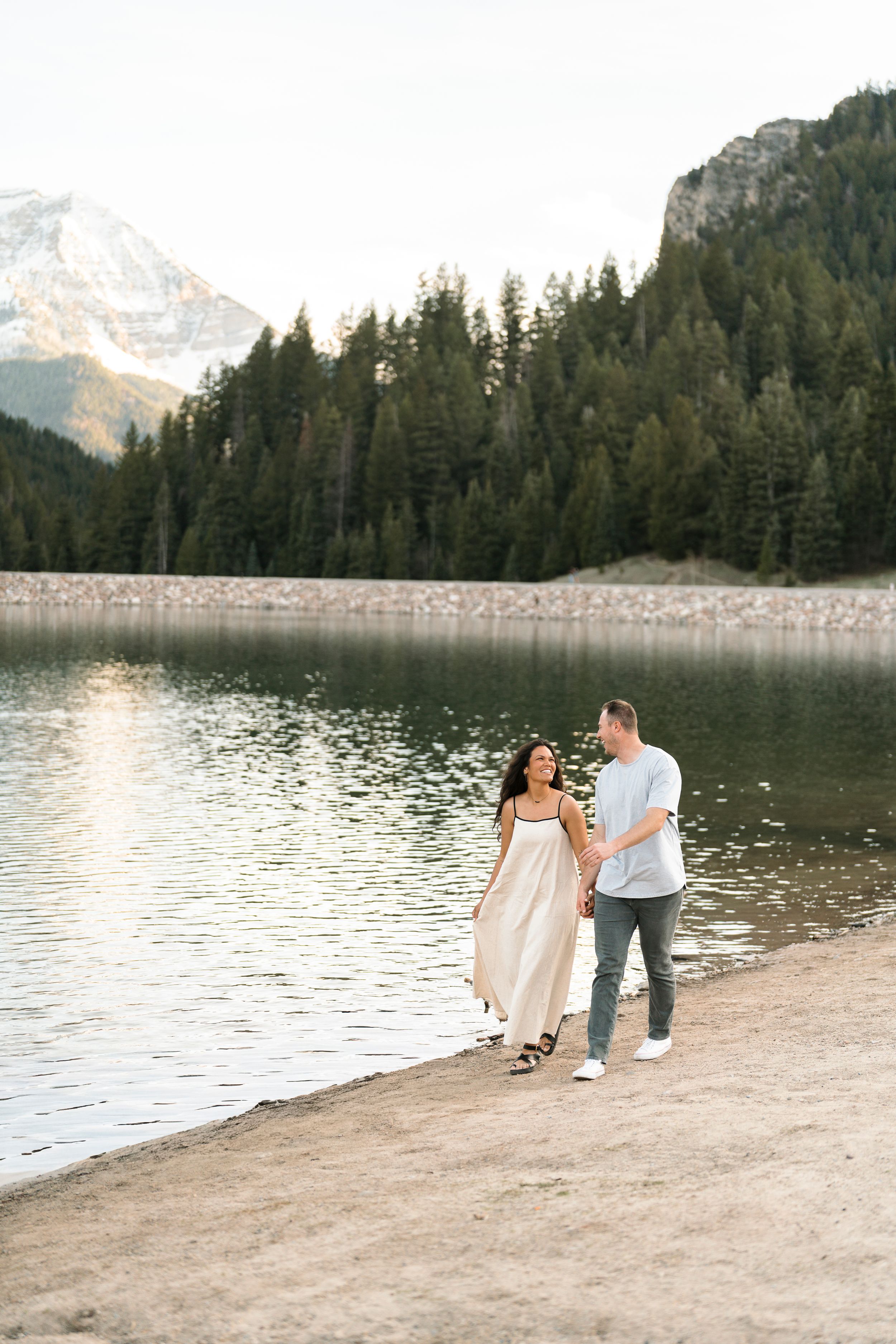Couple walking hand in hand along a mountain lake shore with pine trees and snow-capped peaks in the background.