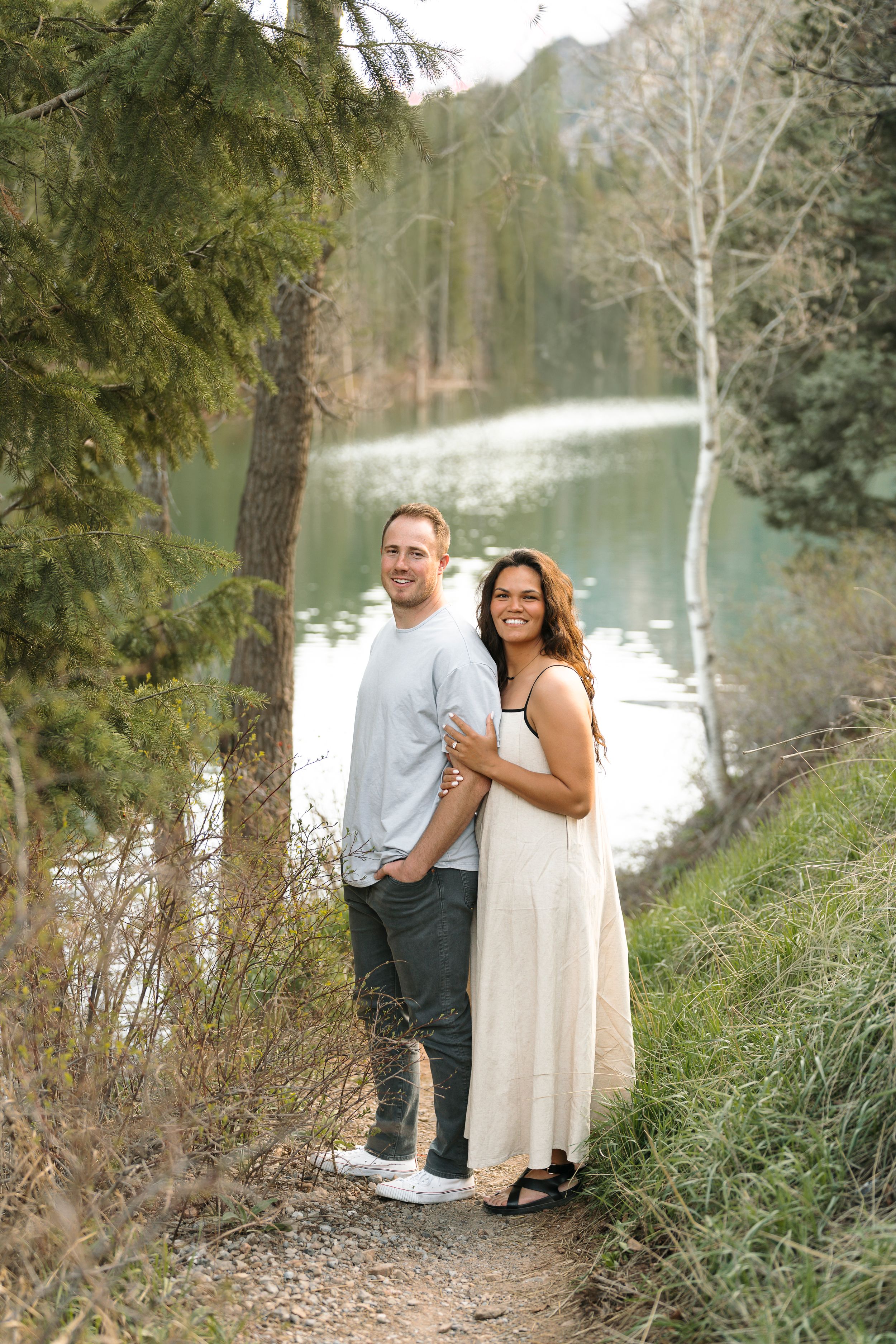 A smiling couple poses together on a lakeside trail surrounded by pine trees and serene water.