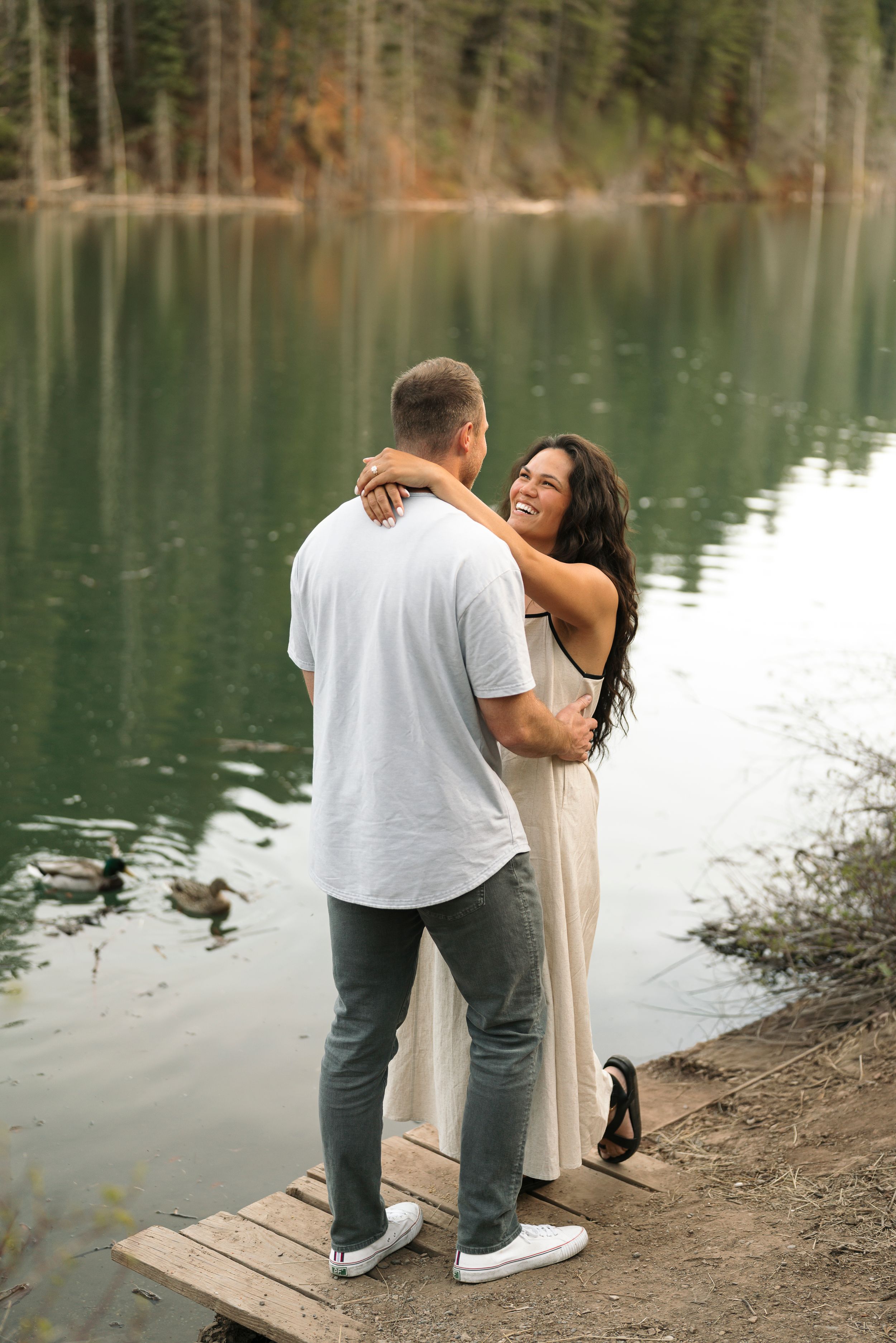 Couple embracing and laughing on a wooden dock by a serene lake surrounded by forest trees.