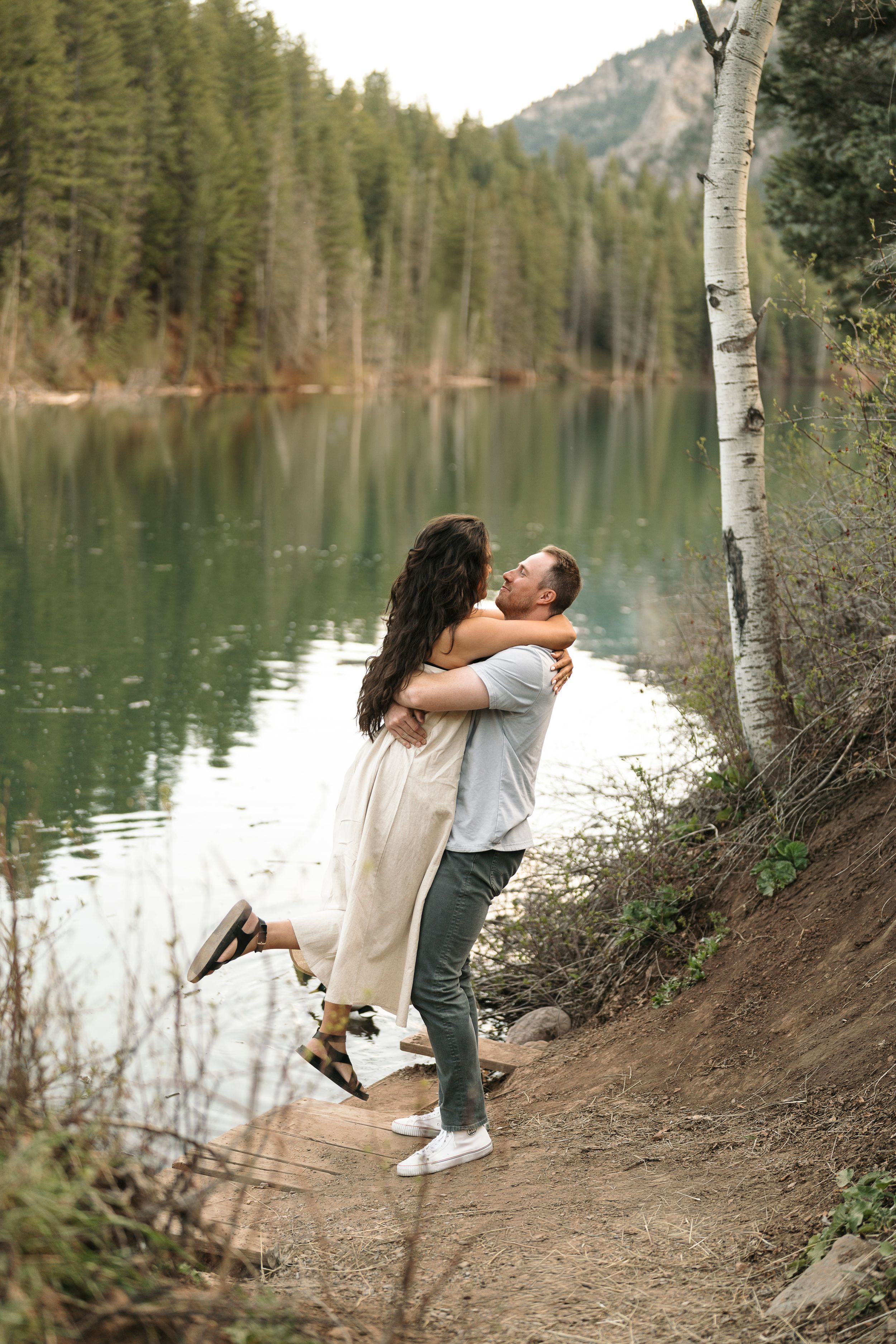 Couple embracing by a serene mountain lake surrounded by evergreen trees and birch, man lifting woman in romantic outdoor setting.