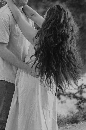 Black and white photo of a couple embracing outdoors, woman with long curly hair in white dress.