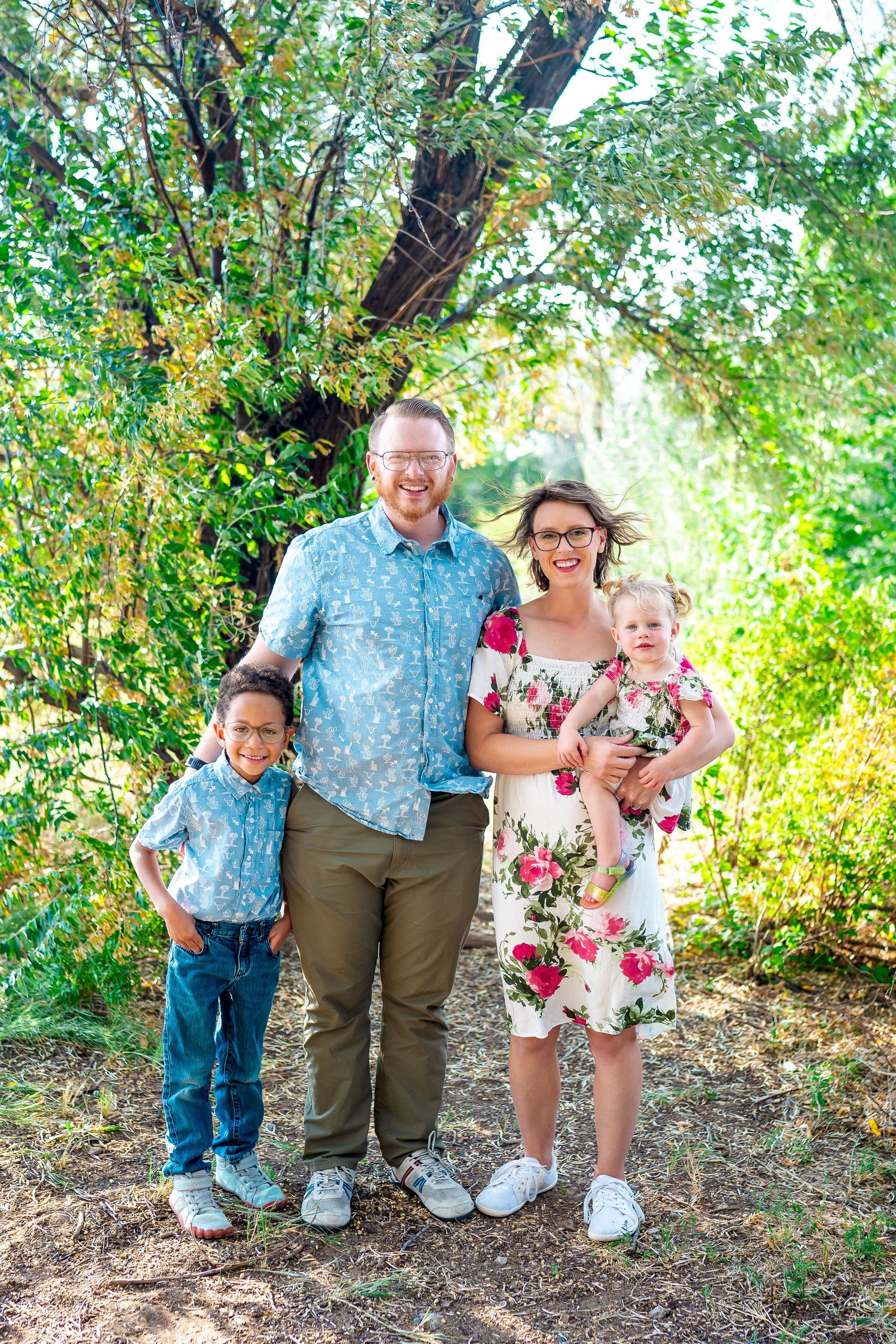 A family of four poses together outdoors in coordinated outfits among green trees.