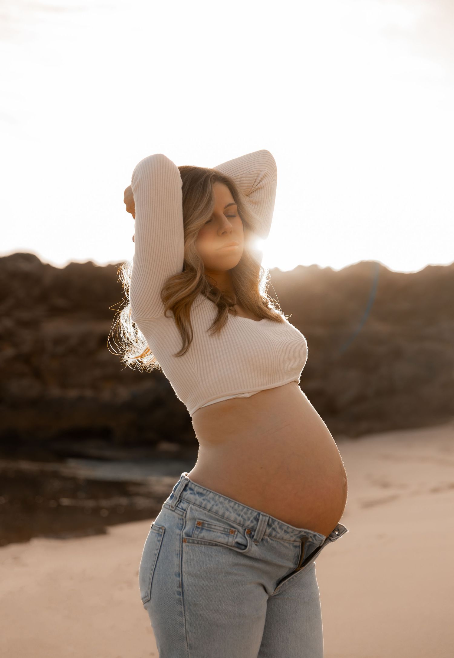 Pregnant woman in cropped sweater and jeans poses at beach during golden hour.