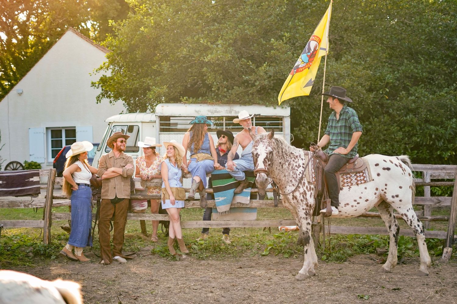 Group of people gathered near a trailer and spotted horse with flag at sunset.
