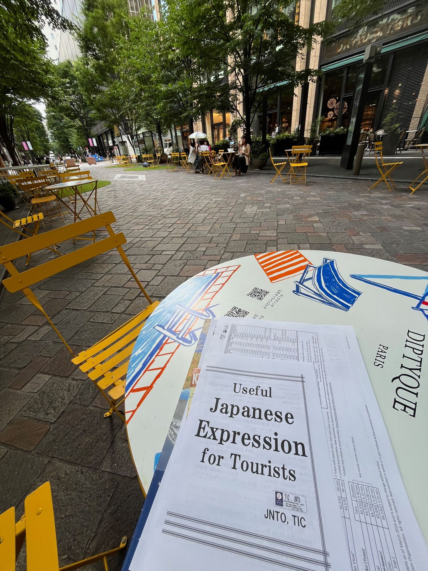 Japanese language guide book on yellow outdoor cafe table along tree-lined street.