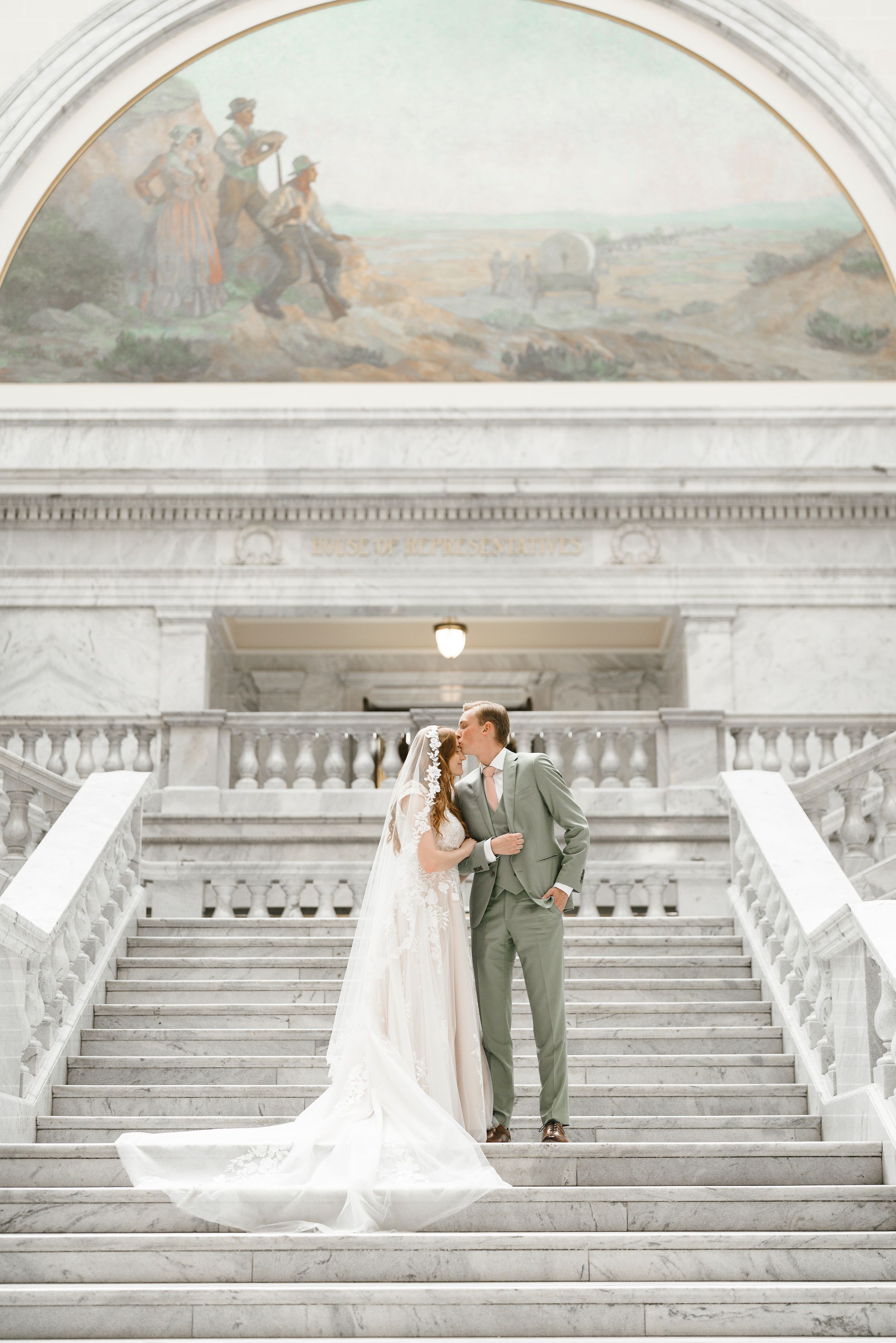 A couple shares a romantic moment on grand marble steps of a historic building with an arched mural above.