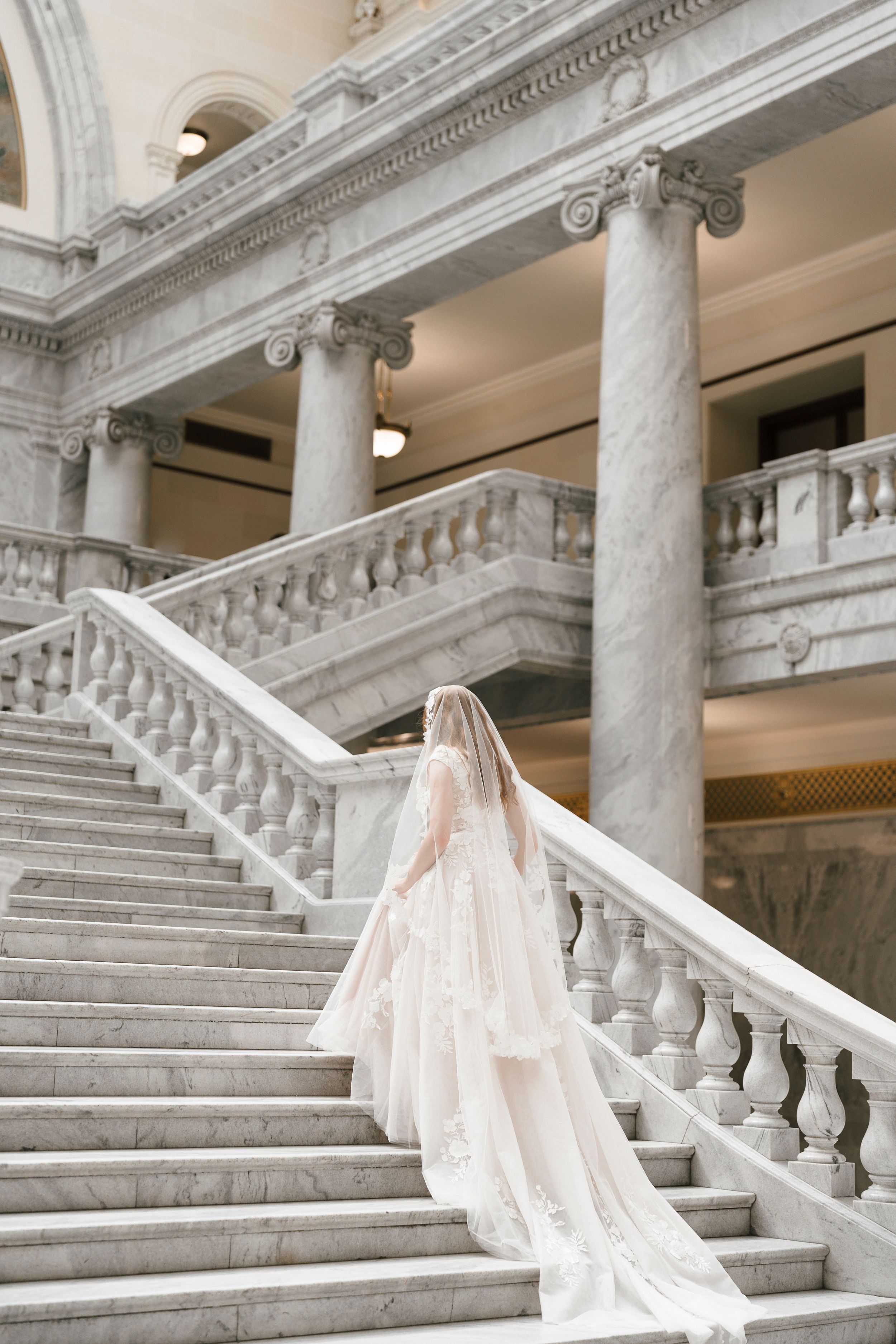 A bride in flowing veil climbs an elegant marble staircase in a historic building with grand columns and ornate details.