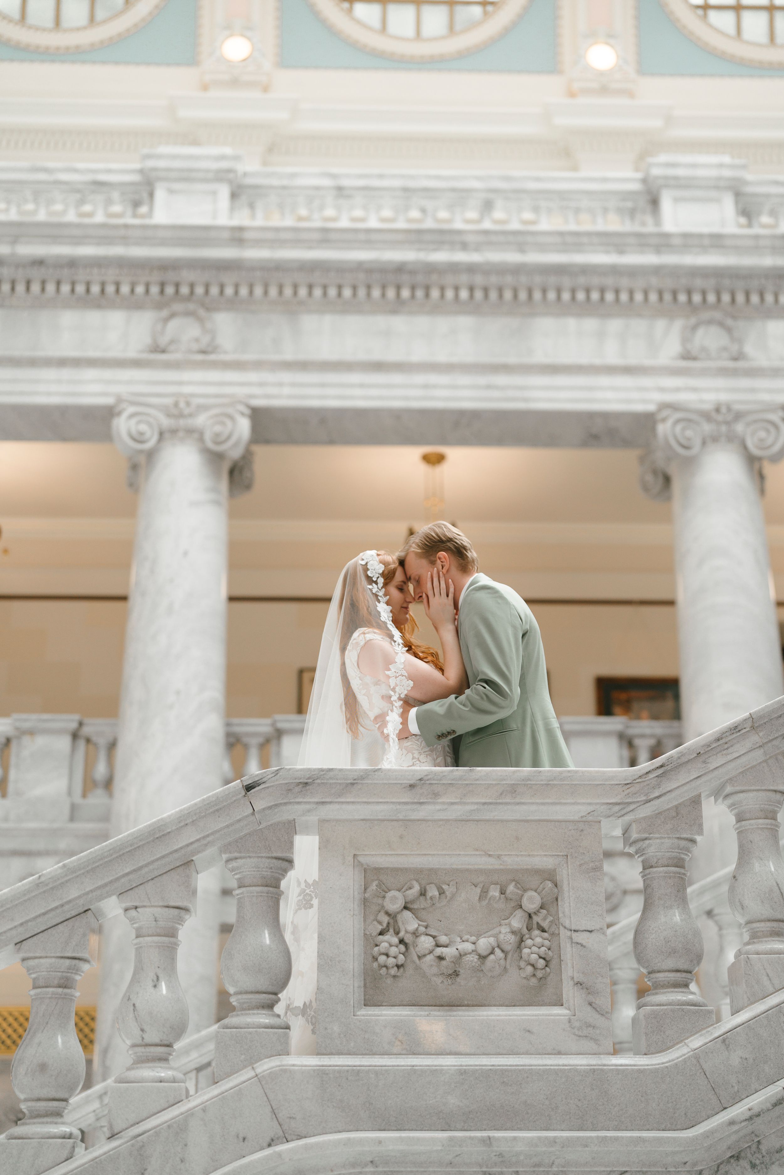 Romantic wedding couple shares intimate moment on ornate marble staircase with white columns and classical architecture.