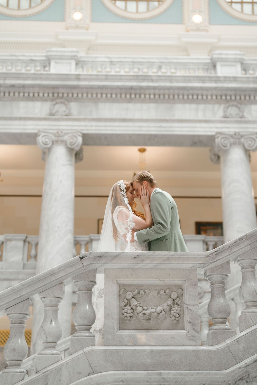 Romantic wedding couple shares intimate moment on ornate marble staircase with white columns and classical architecture.