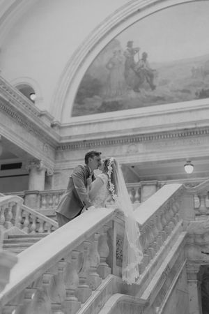 A romantic couple shares an intimate kiss on a grand marble staircase under an ornate arched ceiling with fresco artwork.