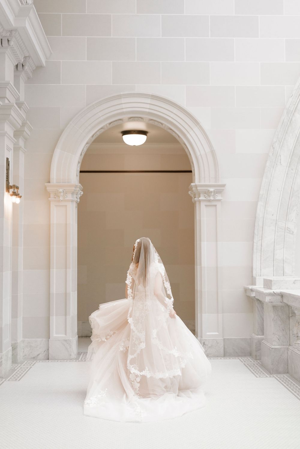 A flowing blush pink wedding gown photographed in an elegant white marble archway with architectural details.