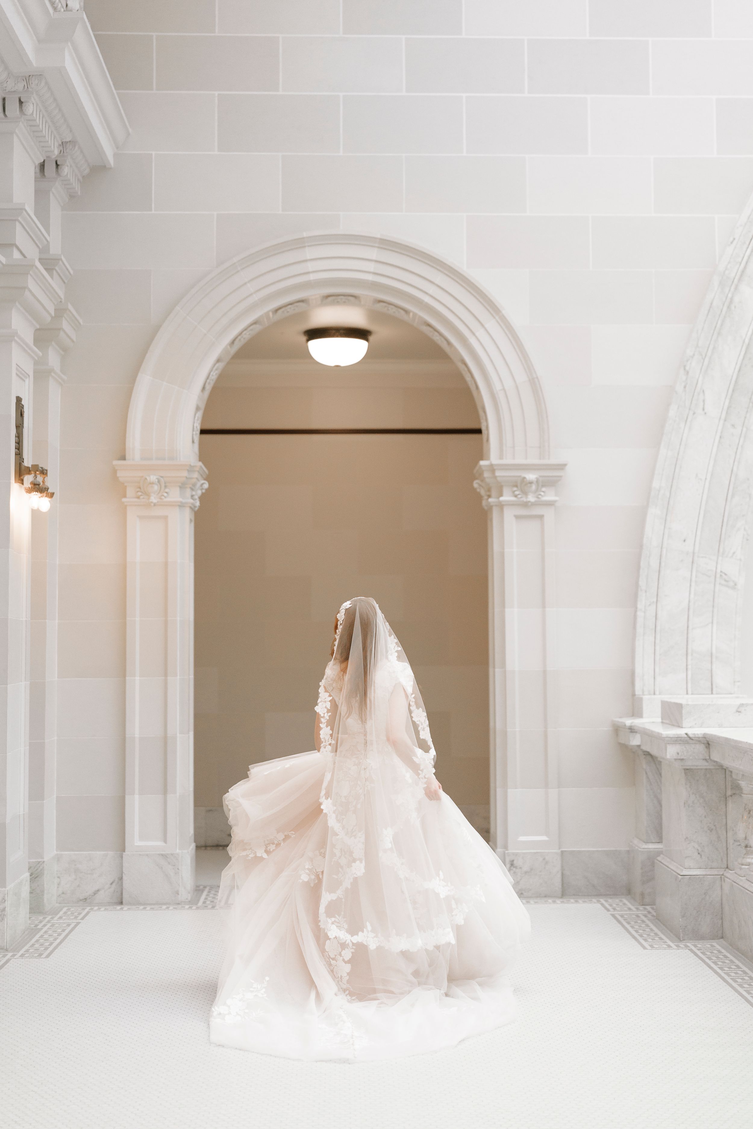 A flowing blush pink wedding gown photographed in an elegant white marble archway with architectural details.