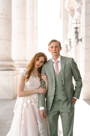 A couple in wedding attire poses elegantly between classical white columns in soft natural lighting.