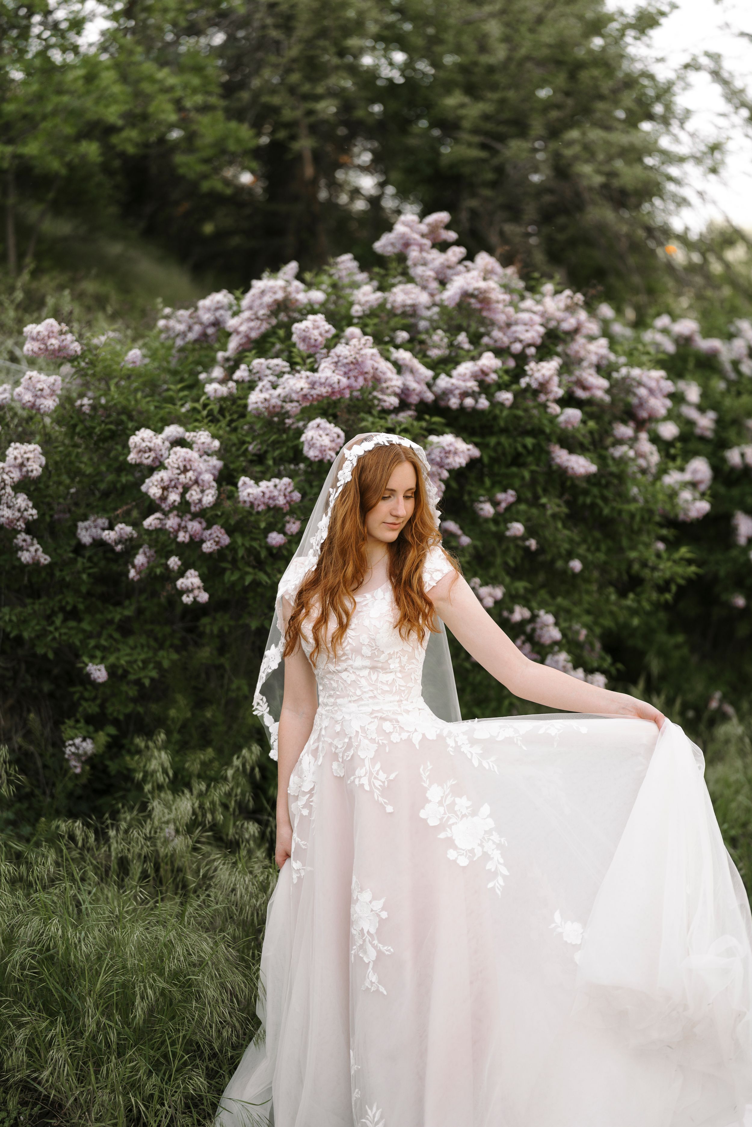 A bride in a delicate lace gown poses gracefully among blooming pink roses in a garden setting.